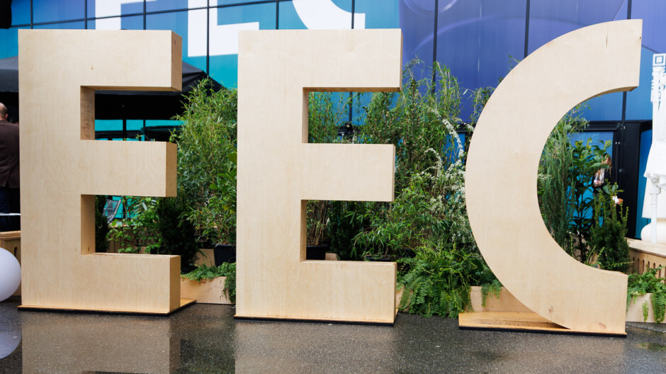 Large wooden letters “EEC” displayed in front of a green plant arrangement outside a building.