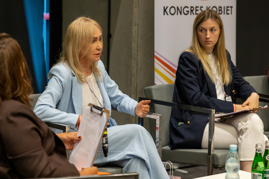 JM Rektor prof. Celina M. Olszak uczestniczy w panelu dyskusyjnym podczas Kongresu Kobiet. Siedzi na fotelu w jasnym garniturze, rozmawia z innymi panelistkami. Obok niej widoczna jest młoda kobieta w ciemnej marynarce.