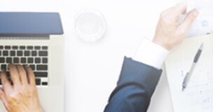 Obrazek przedstawia dłonie kilku osób  trzymających dokumenty oraz laptop, na którym jedna z osób pisze. Całość pokazana jest z góry.