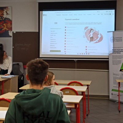 Fotografia przedstawia zajęcia lekcyjne w klasie, podczas których uczniowie oglądają prezentację wyświetlaną na ekranie. Slajd „Czynności zawodowe” zawiera listę zagadnień i ilustrację, a obok stoją materiały informacyjne na stojaku.