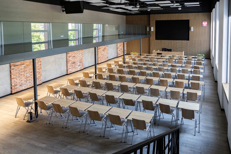 Nowoczesna sala reprezentacyjna na Uniwersytecie Ekonomicznym w Katowicach. Widok na przestronną, jasną aulę z rzędami drewnianych stołów i krzeseł ustawionych w układzie konferencyjnym. Przód sali wyposażony w mównicę, ekran i dekoracyjne panele ścienne. Wnętrze wykończone w industrialnym stylu z elementami cegły, betonu i szkła.