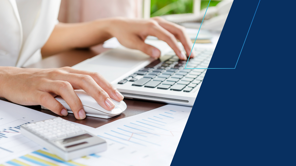 Zdjęcie przedstawia zbliżenie dłoni osoby pracującej przy biurku. Jedna ręka obsługuje mysz komputerową, a druga pisze na klawiaturze laptopa. Na biurku widoczne są dokumenty z wykresami oraz kalkulator, co sugeruje pracę związaną z analizą danych lub finansami.