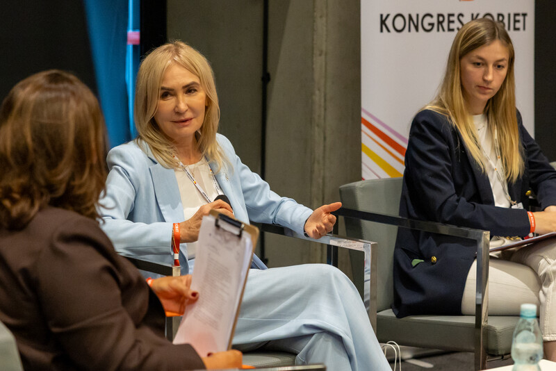 JM Rektor prof. Celina M. Olszak uczestniczy w panelu dyskusyjnym podczas Kongresu Kobiet. Siedzi na fotelu w jasnym garniturze, rozmawia z innymi panelistkami. Obok niej widoczna jest młoda kobieta w ciemnej marynarce.
