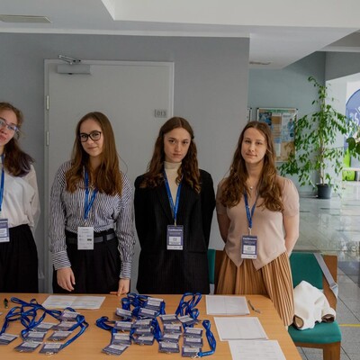 Cztery młode kobiety stojące za stołem rejestracyjnym w holu budynku. Na stole rozłożone są liczne identyfikatory na niebieskich smyczach oraz listy obecności. W tle widać korytarz z roślinami doniczkowymi i wejście do pomieszczenia numer 013.