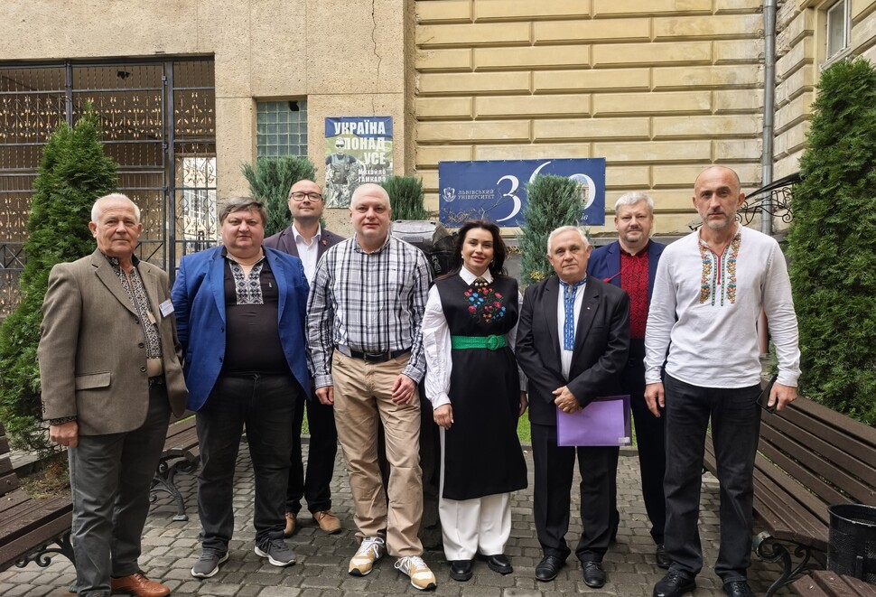 Fotografia przedstawia ośmioosobową grupę naukowców pozującą przed kamiennym budynkiem. W tle widoczne są tablice z napisem „Ukraina” oraz liczbą „30”, a uczestnicy mają tradycyjne ukraińskie hafty.