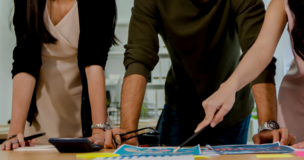 kobieta, mężczyzna oraz kobieta patrzą i wskazują długopisami na kartki z wykresami