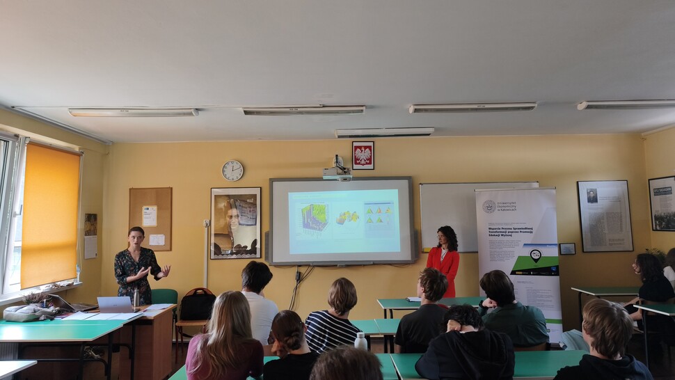 Fotografia przedstawia zajęcia edukacyjne w sali lekcyjnej, podczas których prowadzące prezentują materiały na ekranie projekcyjnym. Uczniowie siedzą przy stołach i słuchają wystąpień; widoczne są tablice informacyjne, polska flaga, zegar oraz kolorowe rolety.