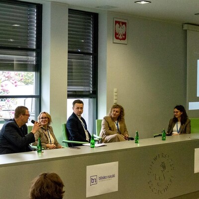 Pięcioro prelegentów siedzących za długim, szarym stołem konferencyjnym podczas panelu dyskusyjnego. Jeden z mężczyzn mówi do mikrofonu. Na przedniej ścianie stołu widoczne są logotypy m.in. Uniwersytetu Szczecińskiego oraz projektów „Menedżer” i „Blisko Nauki”. W tle za oknem widać kwitnące drzewo.
