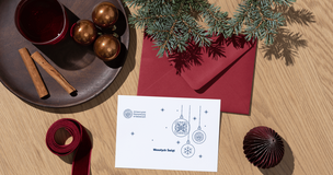 Świąteczna kompozycja na drewnianym blacie: biała kartka z nadrukiem bombek i napisem ‘Wesołych Świąt’ obok bordowej koperty, gałązki świerku, ozdób choinkowych, czerwonej wstążki, talerza z laskami cynamonu oraz szklanki z czerwonym napojem.