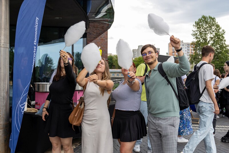 Grupa uśmiechniętych studentów Uniwersytetu Ekonomicznego w Katowicach trzyma watę cukrową podczas plenerowego wydarzenia z okazji Dnia Dziecka. W tle widoczny jest baner uczelni, automat do produkcji waty oraz inni uczestnicy imprezy. Atmosfera jest radosna i swobodna, a studenci celebrują wspólnie czas na świeżym powietrzu.