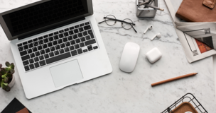 na biurku leży laptop, telefon, słuchawki, okulary, ołówek oraz kwiatek w doniczce