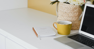 Na biurku leżą laptop, notes, filiżanka i kwiaty