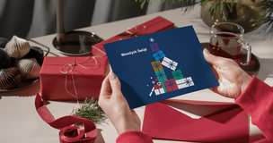 grafika ozdobna: Osoba w czerwonym swetrze trzyma kartkę świąteczną przedstawiającą stylizowaną choinkę z kolorowych prezentów na granatowym tle z napisem „Wesołych Świąt”. W tle na stole znajdują się czerwone prezenty, czerwona koperta, gałązka iglasta, kubek z napojem oraz dekoracje świąteczne.