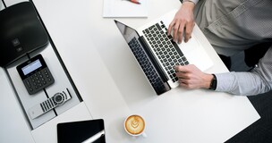 laptop, kubek z kawą i długopisy leżące na biurku