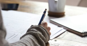 kobieca dłoń trzymająca ołówek i notująca w notesie; w tle kubek