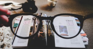 Zbliżenie na okulary trzymane przed otwartym magazynem; przez szkła widać wyraźnie tekst i zdjęcia na stronach, podczas gdy reszta obrazu jest rozmyta. W tle filiżanka i inne przedmioty na stole.