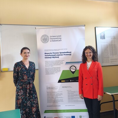Fotografia przedstawia dwie kobiety stojące przy roll-upie Uniwersytetu Ekonomicznego w Katowicach, poświęconym projektowi wspierającemu sprawiedliwą transformację regionu. Plakat zawiera tytuł projektu, opis założeń, grafikę oraz logotypy instytucji i funduszy, a scena znajduje się w sali wykładowej.