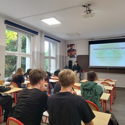Fotografia przedstawia zajęcia edukacyjne w sali lekcyjnej, podczas których prowadząca omawia manipulację danymi. Na ekranie widnieje tytuł „MANIPULACJA DANYMI”, a uczniowie siedzą przy ławkach i słuchają prezentacji; po prawej stronie znajduje się stojak informacyjny.