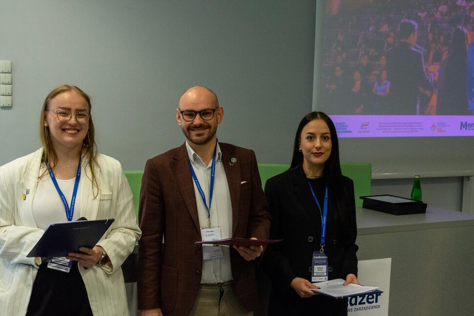 Troje organizatorów – dwie kobiety i mężczyzna – stojących obok siebie i uśmiechających się do aparatu. Trzymają w rękach teczki i dokumenty. Wszyscy mają identyfikatory na niebieskich smyczach Uniwersytetu Szczecińskiego. W tle widać fragment prezentacji multimedialnej.