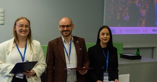 Troje organizatorów – dwie kobiety i mężczyzna – stojących obok siebie i uśmiechających się do aparatu. Trzymają w rękach teczki i dokumenty. Wszyscy mają identyfikatory na niebieskich smyczach Uniwersytetu Szczecińskiego. W tle widać fragment prezentacji multimedialnej.