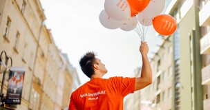 Młody wolontariusz w czerwonej koszulce z napisem ‘WOLONTARIUSZ dkms.pl’ trzyma pęk biało-pomarańczowych balonów z hasłem ‘ZOSTAŃ DAWCĄ SZPIKU’, stojąc na ulicy między budynkami w słoneczny dzień.