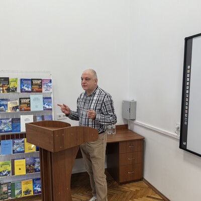 Fotografia przedstawia mężczyznę prowadzącego prezentację przy drewnianej mównicy w sali wykładowej. W tle widoczne są półki z książkami, szafka oraz fragment dużej tablicy interaktywnej, wskazujące na edukacyjny charakter spotkania.