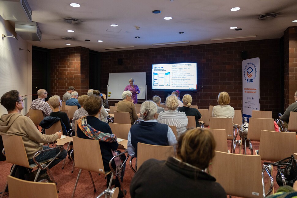 Grupa dorosłych uczestników siedzi na krzesłach w sali wykładowej i słucha prelegentki stojącej przy ekranie. Na ekranie wyświetlana jest prezentacja dotycząca aplikacji mojeIKP.