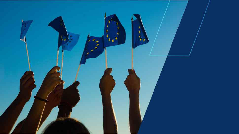 Grupa osób trzyma w górze małe flagi Unii Europejskiej na tle bezchmurnego, niebieskiego nieba; po prawej stronie widoczny jest granatowy, geometryczny panel graficzny.