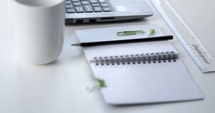 po lewej stronie kubek, po prawej notes, na którym leży długopis, w tle laptop
