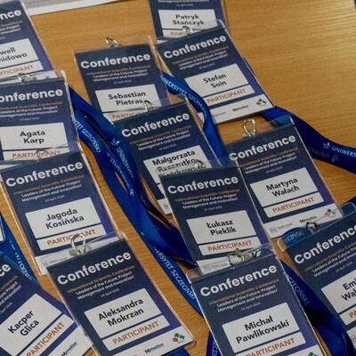 Zbliżenie na dużą grupę identyfikatorów konferencyjnych rozłożonych na drewnianym blacie. Na plakietkach widnieje napis „Conference”, data „24 April 2026” oraz imiona i nazwiska uczestników