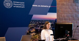wykładowczyni siedzi w sali wykładowej przy biurku z komputerem, uczestniczy w spotkaniu online