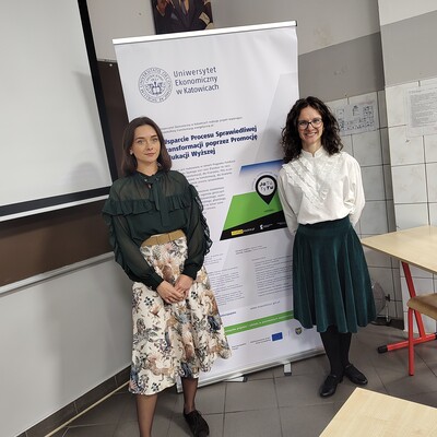 Fotografia dokumentuje spotkanie dwóch kobiet w sali szkolnej, stojących obok roll-upu promującego projekt wsparcia procesu sprawiedliwej transformacji poprzez promocję edukacji wyższej. Baner zawiera logo uczelni, tytuł projektu, grafikę oraz oznaczenia instytucji finansujących, a w tle widoczne są ekran projekcyjny i stoliki szkolne.