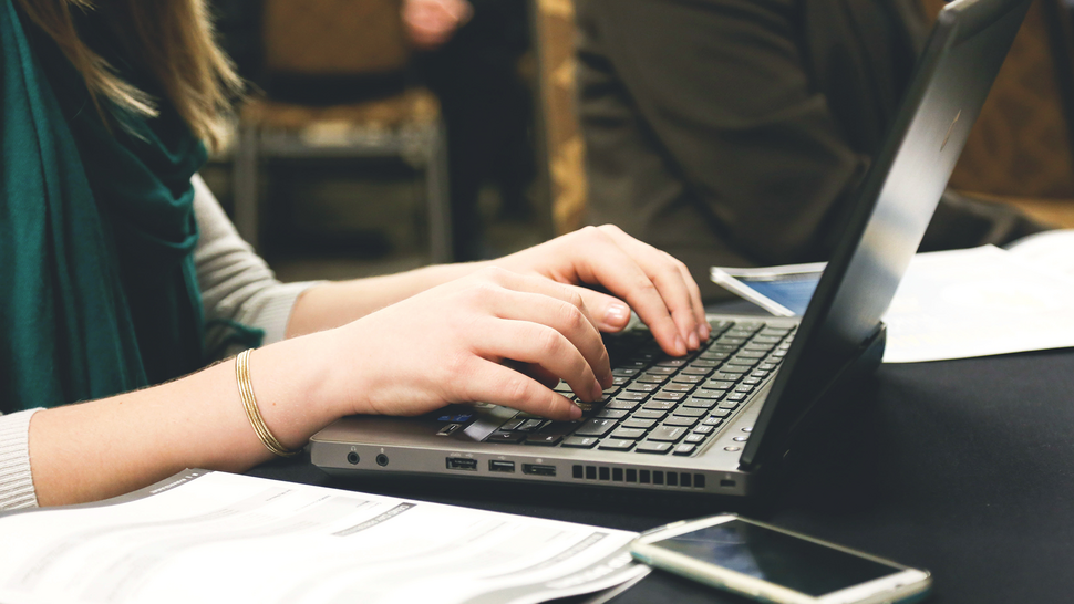 widok rąk piszących na laptopie, na pierwszym planie widać kartki i telefon komórkowy