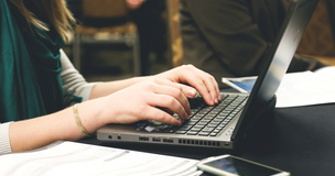 widok rąk piszących na laptopie, na pierwszym planie widać kartki i telefon komórkowy