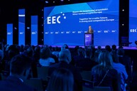 Prezes Zarządu PTWP SA Wojciech Kuśpik otwiera Europejski Kongres Gospodarczy (EEC). Stoi przy mównicy na tle dużego, niebieskiego ekranu z hasłem wydarzenia w języku polskim i angielskim: „Razem dla bezpiecznej przyszłości. Silna i konkurencyjna Europa / Together for a safer future. A strong and competitive Europe”. Przed sceną zgromadzona jest licznie publiczność. Wystąpienie odbywa się w nowoczesnej, profesjonalnej oprawie audiowizualnej.