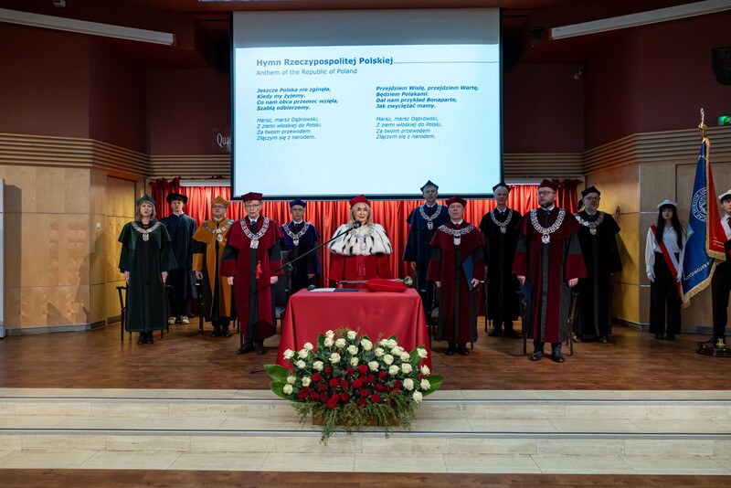 JM Rektor, Prorektorzy oraz Władze Wydziałów na scenie podczas uroczystości, w tle slajd z hymnem narodowym