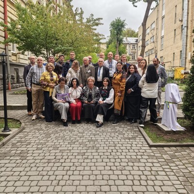 Fotografia grupowa przedstawiająca około 25 osób pozujących na brukowanej uliczce między zabytkowymi kamienicami. W centrum stoją i siedzą uczestnicy w różnorodnych strojach, a otoczenie tworzą drzewa, latarnie, ławki oraz jasne budynki miejskie.