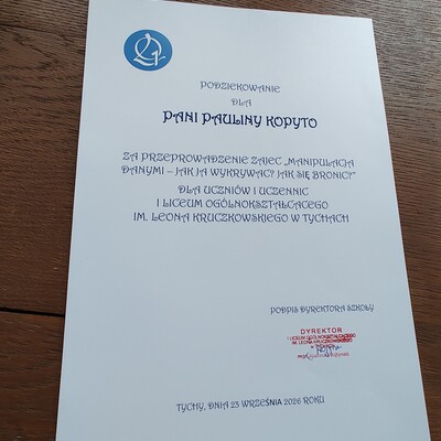 Fotografia podziękowania dla prowadzącej za przeprowadzenie zajęć dla uczniów i uczennic dotyczących bezpieczeństwa w sieci oraz cyberobrony. Biała karta z niebieskim logo, wyśrodkowanym tekstem, podpisem i pieczęcią u dołu oraz datą 25 września 2026 roku, leżąca na drewnianym stole.