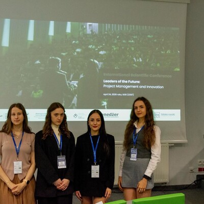Cztery młode kobiety stojące w rzędzie na tle ekranu projekcyjnego w sali konferencyjnej. Wszystkie mają na szyjach niebieskie smycze z identyfikatorami. Na ekranie za nimi widnieje napis: „International Scientific Conference. Leaders of the Future: Project Management and Innovation. April 24, 2026”.