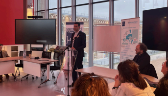 Speaker presenting at an academic event in a modern conference room with audience and presentation screen.